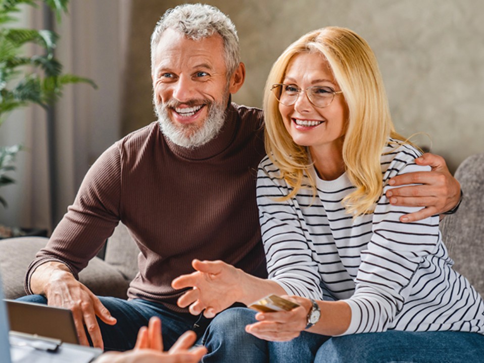 Man and woman are sitting at the desk of a personal banker. The Banker has their back to the camera. Man and woman are smiling.